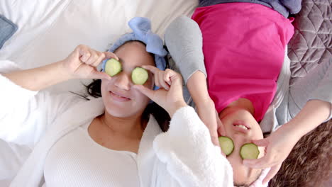 Happy-biracial-mother-and-daughter-lying-on-bed-with-cucumber-slices-on-eyes