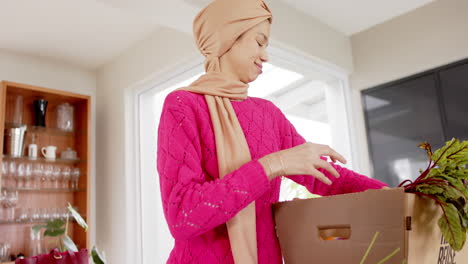 Feliz-Mujer-Birracial-En-Hijab-Con-Compras-En-La-Cocina-De-Casa-Con-Espacio-Para-Copiar,-Cámara-Lenta