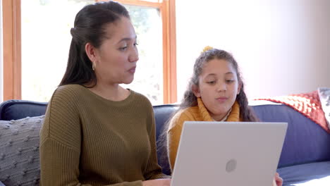 Happy-biracial-mother-and-daughter-sitting-on-sofa,-using-laptop-and-talking-in-sunny-living-room