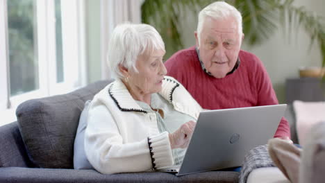 Happy-senior-caucasian-couple-sitting-on-sofa-using-laptop-at-home,-slow-motion
