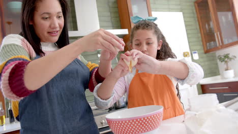 Happy-biracial-mother-and-daughter-preparing-dough-and-smiling-in-sunny-kitchen