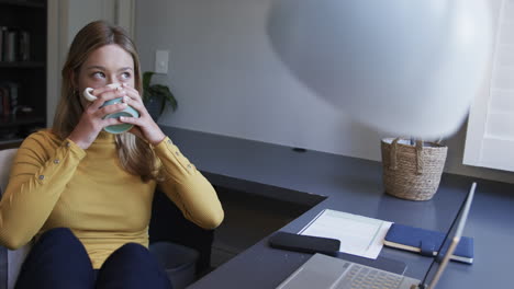 Biracial-woman-sitting-at-desk-using-laptop-and-drinking-coffee-at-home,-slow-motion