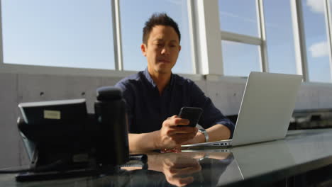 Animation-of-multiple-icons-over-asian-man-sitting-on-chair-and-scrolling-on-cellphone-in-office