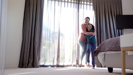 Happy-biracial-mother-and-daughter-dancing-and-embracing-in-sunny-bedroom