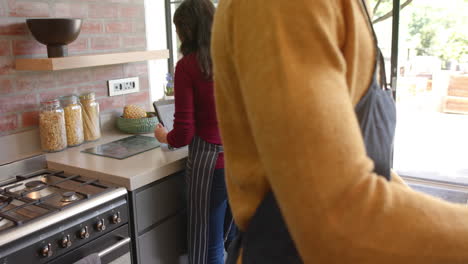 Happy-diverse-couple-in-aprons-using-tablet-and-baking-in-sunny-kitchen,-slow-motion