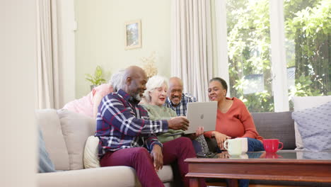 Fröhliche,-Vielfältige-Seniorenfreunde,-Die-Gemeinsam-Einen-Laptop-Im-Sonnigen-Wohnzimmer-Benutzen,-Platz-Zum-Kopieren,-Zeitlupe