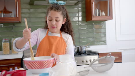 Happy-biracial-girl-with-long,-curly-hair-mixing-dough-in-bowl-and-smiling-in-sunny-kitchen