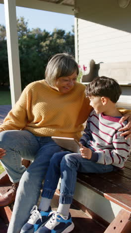 Vertical-video-of-biracial-grandmother-and-grandson-sitting-on-terrace-using-tablet,-slow-motion