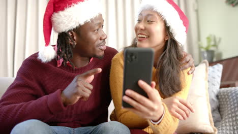 Happy-diverse-couple-in-christmas-hats-using-smartphone-talking-on-couch-at-home,-slow-motion