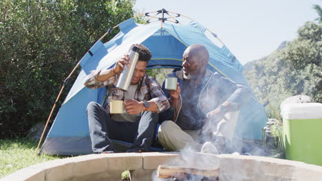 Happy-african-american-father-and-adult-son-sitting-outside-tent-drinking-coffee-in-sun,-slow-motion