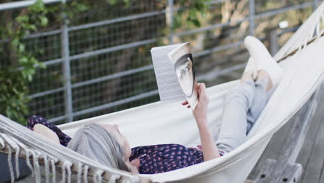 Caucasian-middle-aged-woman-reading-book-relaxing-in-hammock-on-terrace-in-nature,-slow-motion