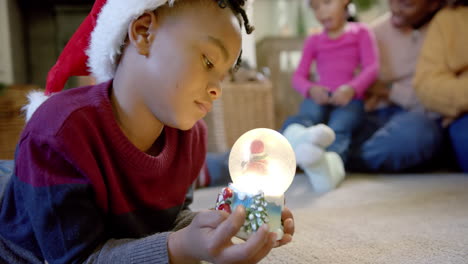 African-american-son-in-christmas-hat-holding-glowing-snow-globe,-family-in-background,-slow-motion