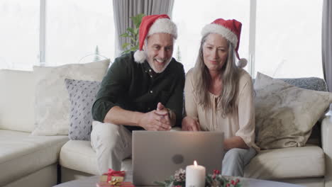 Middle-aged-caucasian-couple-in-santa-hats-on-laptop-video-chat-at-christmas-at-home,-slow-motion