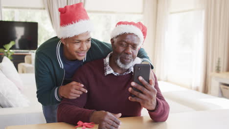 Feliz-Padre-E-Hijo-Afroamericanos-Con-Sombreros-Navideños-Teniendo-Videollamadas-Con-Teléfonos-Inteligentes,-Cámara-Lenta