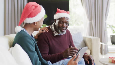 Glücklicher-Afroamerikanischer-Vater-Und-Erwachsener-Sohn-In-Weihnachtsmützen-Mit-Tablet-Videoanruf,-Zeitlupe