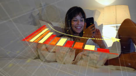 Animation-of-arrow-on-falling-graph,-smiling-biracial-woman-lying-on-bed-and-scrolling-on-cellphone