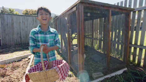 Portrait-of-happy-biracial-boy-holding-basket-with-eggs-next-to-henhouse,-slow-motion,-copy-space