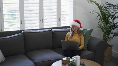 Happy-biracial-woman-wearing-santa-hat-using-laptop-at-christmas,-copy-space,-slow-motion
