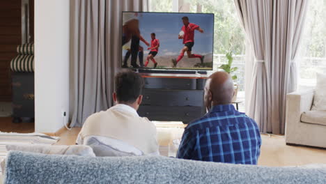 Rear-view-of-african-american-father-and-adult-son-talking-and-watching-rugby-on-tv,-slow-motion