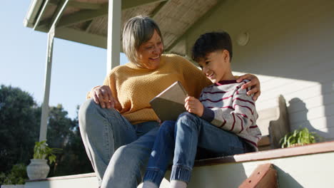 Happy-senior-biracial-grandmother-and-grandson-using-tablet-sitting-on-terrace,-slow-motion