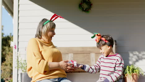 Happy-biracial-grandmother-and-grandson-sitting-on-terrace-giving-christmas-presents,-slow-motion