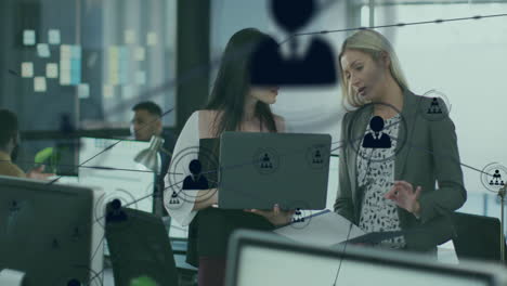 Animation-of-connected-icons-over-diverse-female-coworkers-standing-and-discussing-reports-on-laptop