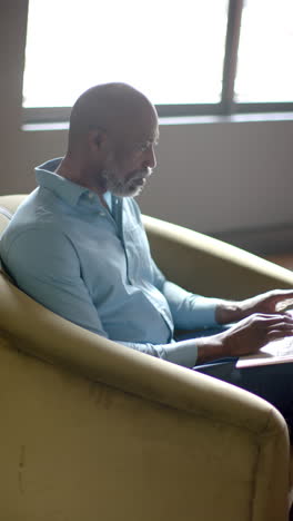 Vertical-video-of-african-american-businessman-using-laptop-in-office-with-copy-space