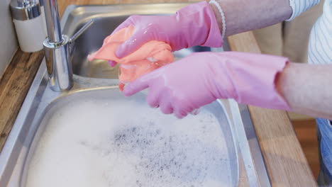 Middle-aged-hands-of-caucasian-woman-washing-up-in-gloves-in-kitchen-with-copy-space