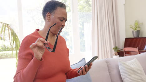 Glückliche-ältere-Afroamerikanische-Frau-Mit-Brille,-Die-Ihr-Smartphone-Im-Sonnigen-Wohnzimmer-Benutzt,-Zeitlupe