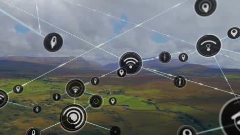Animation-of-connected-icons-over-aerial-view-of-green-landscape-against-mountains-and-cloudy-sky