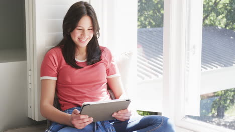 Happy-biracial-woman-sitting-by-window-in-sunny-room-using-tablet,-copy-space,-slow-motion