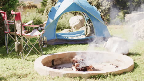 Smoking-firepit,-tent,-chairs-and-camping-equipment-in-sunny-countryside,-copy-space,-slow-motion