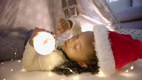Happy-african-american-girl-in-christmas-hat-holding-glowing-snow-globe-in-blanket-tent,-slow-motion