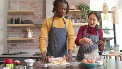 Happy-diverse-couple-in-aprons-using-tablet-and-baking-in-kitchen,-slow-motion