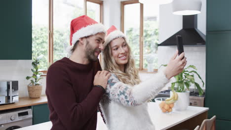 Diverse-couple-wearing-santa-hats-using-smartphone-and-taking-selfies,-in-slow-motion