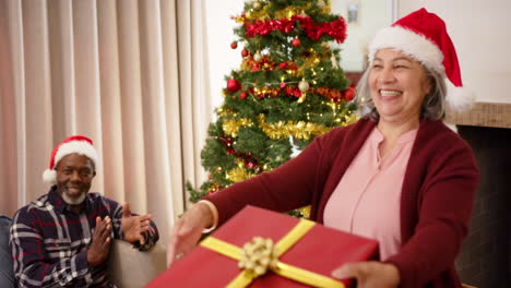 Happy-diverse-senior-couple-in-christmas-hats-exchanging-gift-and-embracing-at-home,-slow-motion