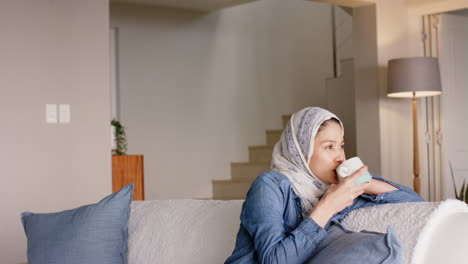 Happy-biracial-woman-in-hijab-drinking-tea-on-sofa-at-home-with-copy-space,-slow-motion