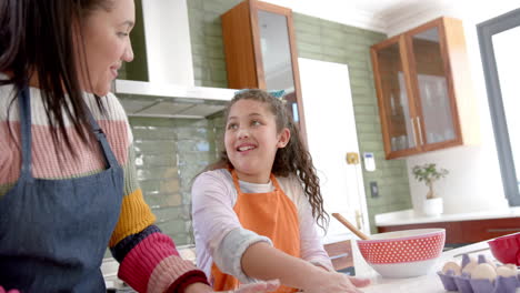Happy-biracial-mother-and-daughter-putting-dough-into-cake-tin-and-smiling-in-sunny-kitchen