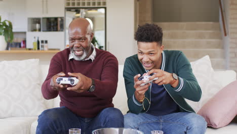 Aufgeregter-Afroamerikanischer-Vater-Und-Erwachsener-Sohn-Spielen-Zu-Hause-Videospiele-Mit-Gamepads,-Zeitlupe