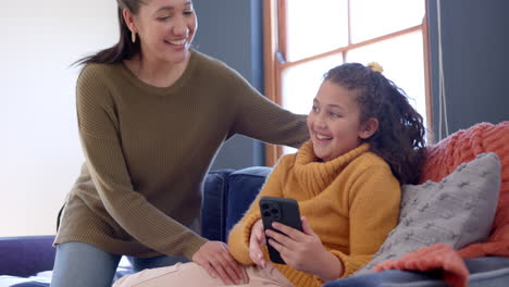 Happy-biracial-mother-and-daughter-embracing-on-sofa-and-using-smartphone-in-sunny-living-room