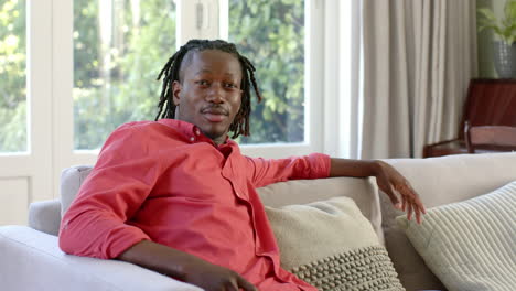 Portrait-of-happy-african-american-man-with-dreadlocks-smiling-on-couch-in-sunny-room,-slow-motion