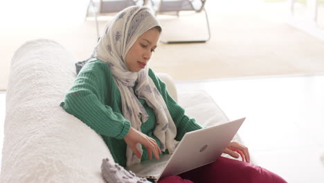 Biracial-woman-in-hijab-using-laptop-on-sofa-at-home-with-copy-space,-slow-motion