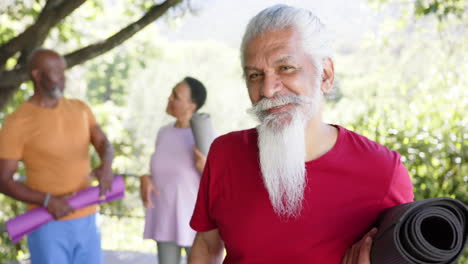 Portrait-of-happy-biracial-man-with-white-beard-and-yoga-mat-out-in-sun-with-friends,-slow-motion