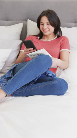 Vertikales-Video-Einer-Glücklichen,-Gemischtrassigen-Frau,-Die-Auf-Dem-Bett-Sitzt,-Ein-Buch-Liest-Und-Ihr-Smartphone-Benutzt,-Zeitlupe