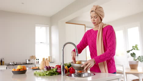 Biracial-woman-in-hijab-washing-food-in-kitchen-at-home-with-copy-space,-slow-motion