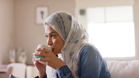 Mujer-Birracial-Feliz-Con-Hijab-Bebiendo-Té-En-El-Sofá-De-Casa-Con-Espacio-Para-Copiar,-Cámara-Lenta