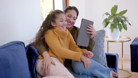 Happy-biracial-mother-and-daughter-embracing-on-sofa-and-using-tablet-in-sunny-living-room