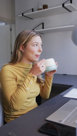 Vertical-video-of-portrait-of-happy-biracial-woman-drinking-coffee-at-desk-at-home,-slow-motion
