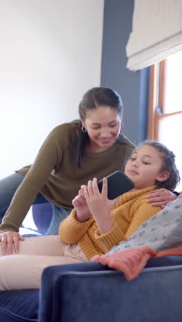 Glückliche-Biracial-Mutter-Und-Tochter-Sitzen-Auf-Dem-Sofa-Und-Mit-Smartphone-Im-Sonnigen-Wohnzimmer