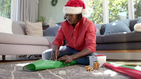 Happy-african-american-man-in-christmas-hat-wrapping-gift-on-floor-at-home,-slow-motion